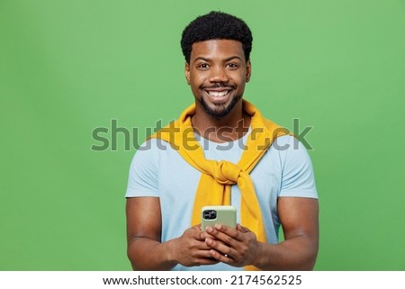 Young man of African American ethnicity 20s wear blue t-shirt hold in hand use mobile cell phone chatting browsing internet isolated on plain green background studio portrait. People lifestyle concept