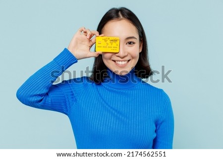 Fascinating fun joyful young woman of Asian ethnicity 20s years old wears blue shirt hold in hand credit bank card cover close hiding eye isolated on plain pastel light blue background studio portrait Royalty-Free Stock Photo #2174562551