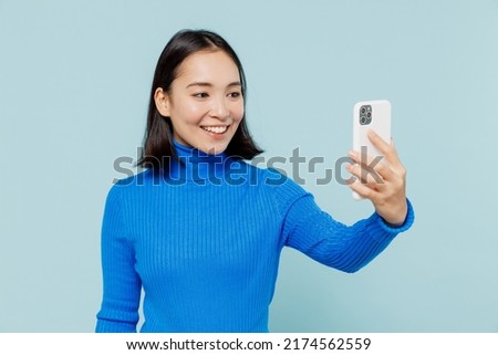 Smiling young woman of Asian ethnicity 20s years old wears blue shirt doing selfie shot on mobile cell phone post photo on social network isolated on plain pastel light blue background studio portrait
