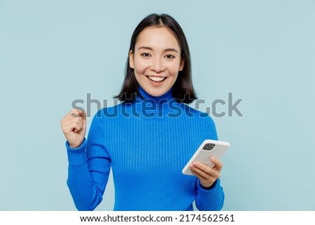 Fun young woman of Asian ethnicity 20s years old wears blue shirt hold in hand use mobile cell phone doing winner gesture clenching fists isolated on plain pastel light blue background studio portrait Royalty-Free Stock Photo #2174562561