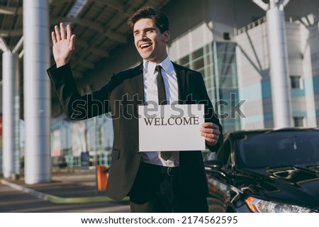 Bottom view young friendly traveler businessman man in black suit stand outside at international airport terminal hold card sign with welcome title text waving hand Air flight business trip concept.