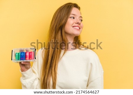 Young caucasian woman holding a battery box isolated on yellow background looks aside smiling, cheerful and pleasant.