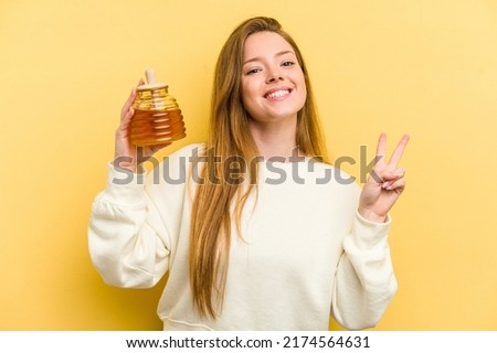 Young caucasian woman holding a honey isolated on yellow background joyful and carefree showing a peace symbol with fingers.