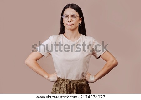 Waist-up portrait of beautiful girl with brunette straight hair in displeasure, wears beige t-shirt, eyeglasses, poses with hands on the waist. Attractive young angry and upset woman in closed posture