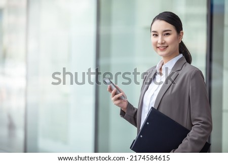 Asian woman with smartphone standing against street blurred building background, asian business photo of beautiful girl in casual suite with smart phone.  Royalty-Free Stock Photo #2174584615