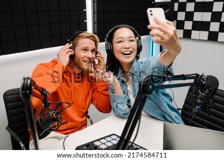 Happy young man and woman smiling and taking selfie photo while broadcasting in radio studio
