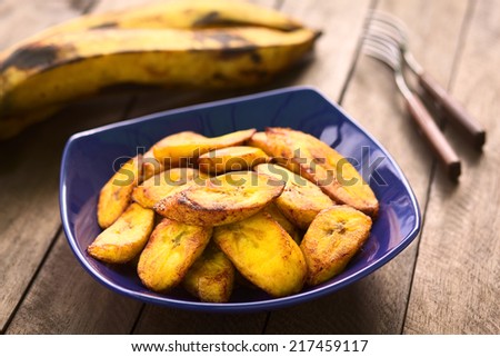 Fried slices of the ripe plantain in blue bowl, which are eaten as snack or used to accompany dishes in some South American countries (Selective Focus, Focus on the front of the upper plantain slice) Royalty-Free Stock Photo #217459117
