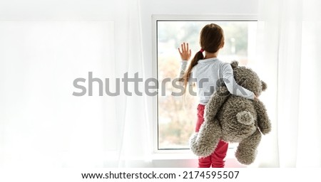 Loneliness. Lonely orphan girl in an orphanage looking outside while holding teddy bear the window. Child feelings concept Royalty-Free Stock Photo #2174595507