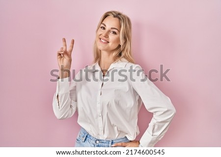 Young caucasian woman wearing casual white shirt over pink background smiling looking to the camera showing fingers doing victory sign. number two. 