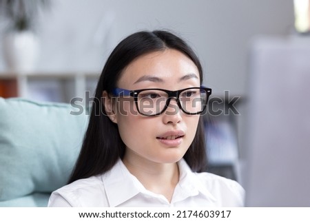 Close-up photo portrait of a young Asian woman office worker, wearing glasses and a white shirt, working in the office, looking at the monitor.