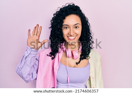 Young hispanic woman with curly hair wearing gym clothes and using headphones showing and pointing up with fingers number five while smiling confident and happy. 