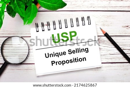 On a light wooden background, a magnifying glass, a pencil, a green plant and a white notebook with text USP Unique Selling Proposition. Business concept
