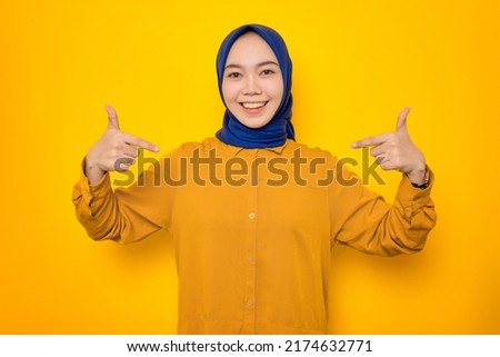 Smiling young Asian Muslim woman dressed in orange shirt pointing at herself with proud isolated on yellow background