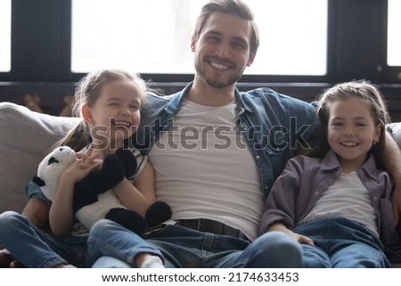 Little 5s 8s daughters sitting next to daddy at home smile look at camera. Loving dad hugging pretty kids pose for picture, capture moment for family album. People celebrating Happy Father Day concept