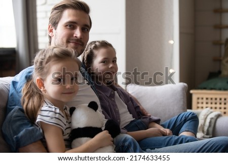 Little 5s 8s daughters sitting next to daddy at home smile look at camera. Loving dad hugging pretty kids pose for picture, capture moment for family album. People celebrating Happy Father Day concept