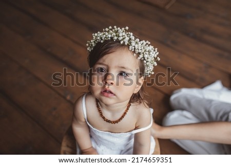 Portrait of pretty one year girl in white summer dress, flower crown and with eco beads sitting in the studio near mom's legs. Eco style concept Royalty-Free Stock Photo #2174659351