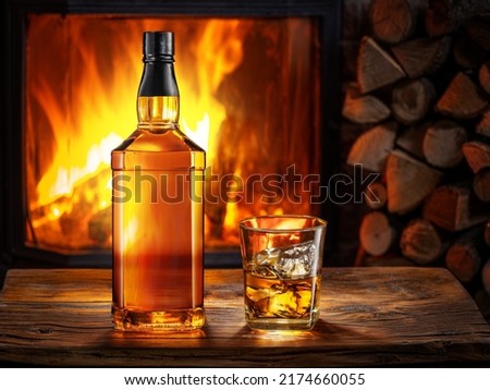 Bottle of whiskey and glass of whiskey with ice. Burning fire in the fireplace at the background.