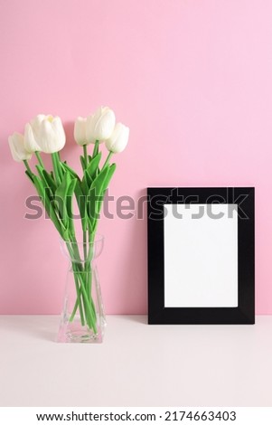 Home decoration with floral decor, frame poster on table. Beautiful flowers white tulips in vase on pink background. 