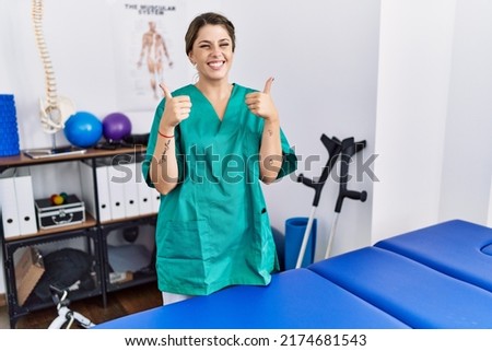Young hispanic woman wearing physiotherapist uniform standing at clinic success sign doing positive gesture with hand, thumbs up smiling and happy. cheerful expression and winner gesture. 