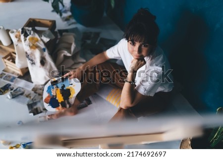 Pensive young female artist draws  picture with paints and enjoys relaxing process of her hobby.  Creative and talented brunette woman creates picture on easel. Learning to draw with oil