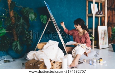 Excited young female artist draws  picture with paints and enjoys relaxing process of her hobby.  Creative happy brunette woman creates picture on easel in studio. Learning to draw with oil