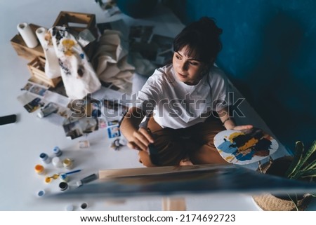Attractive young female artist draws  picture with paints and enjoys relaxing process of her hobby.  Creative and talented brunette woman creates picture on easel. Learning to draw with oil Royalty-Free Stock Photo #2174692723
