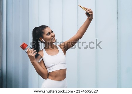 Attractive healthy female in earphones photographing herself on smartphone camera, standing on copy space promo background. Young smiling woman blogger with fit figure posing for video on cellphone