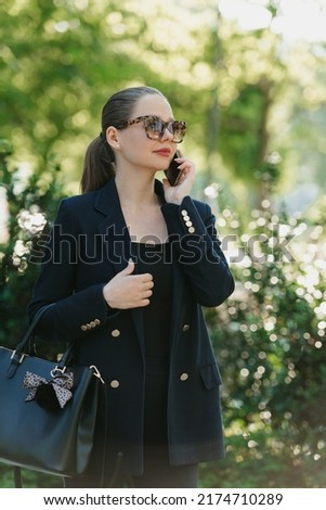 A photo from the side of a female realtor in a blazer who is having a conversation on a phone in the modern garden at sunset in the financial district. Businesswoman in the contemporary urban space.