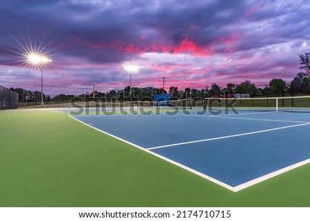 Evening photo of new outdoor blue tennis courts with green out of bounds area and light blue pickleball lines with lights turned on.	