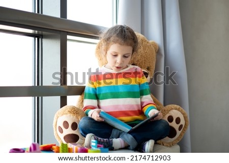 A cute girl of 6 years old sits on the couch by the window with a large toy bear and reads a book. She is dressed in a colored jumper. Kids. Teaching. Royalty-Free Stock Photo #2174712903