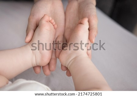 Close up shot loving mom palms holding feet, tiny toes of new born baby. Unrecognizable mother touches barefoot legs of 0-6 child, enjoy bonding, moments with infant feeling unconditional love concept