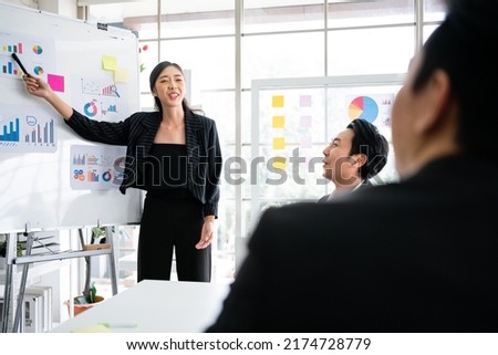 A cheerful and confident Asian businesswoman stands, present and point with a pen to bar charts data from a whiteboard to her office colleagues. Asian business women leader role at the meeting.
