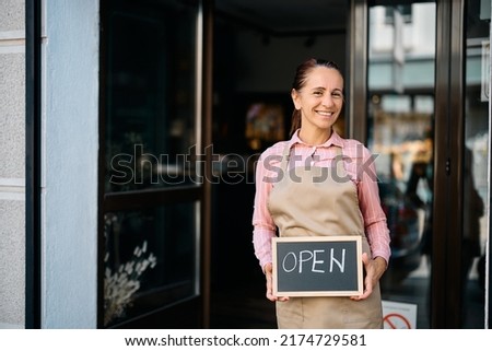 Happy florist holding open sign at entrance door of her flower shop.