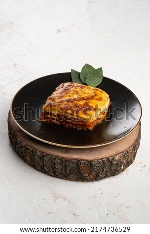 This is picture of Lasagna on a wooden base with black plate and white background.