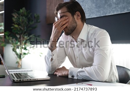 Tired young man feel pain eyestrain holding glasses rubbing dry irritated eyes fatigued from computer work, stressed man suffer from headache bad vision sight problem sit at office Royalty-Free Stock Photo #2174737893