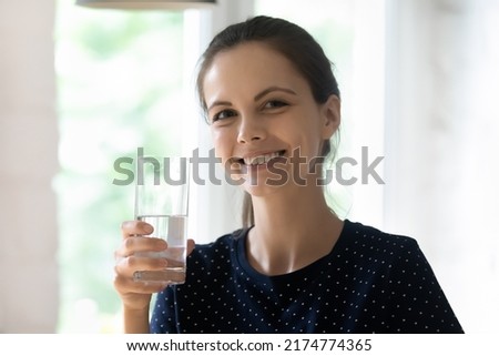 Happy cheerful beautiful girl holding clear transparent glass of fresh pure drink water indoors, looking at camera smiling. Home head shot portrait. Healthy habit, routine concept