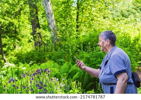 Senior woman taking picture with mobile phone of beautiful blue flowers in summer forest park. Female tourist uses cell phone, technology for social media. forest and blooming flowers on background