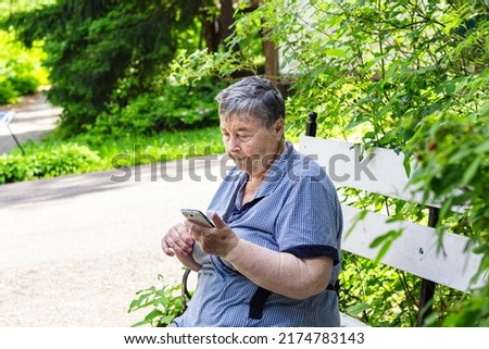 female posing against amazing background, sitting on bench, surrounded with green bushes and flowers. Senior woman uses cell phone to call or chat.Elderly community concept.grandparents day.