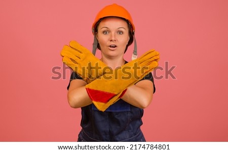 woman in protective overalls, gloves and a helmet shows a stop sign with crossed arms on a pink background