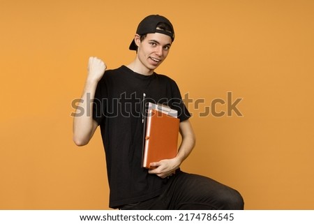 Joyful young guy student in a black baseball cap with notebooks in his hands on a yellow background with copy space