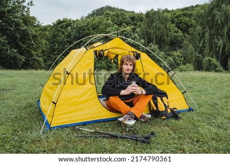 A guy sitting in a campsite in a tent holding a mug of hot tea, hiking in the forest, trekking in the mountains, a summer vacation in nature. High quality photo