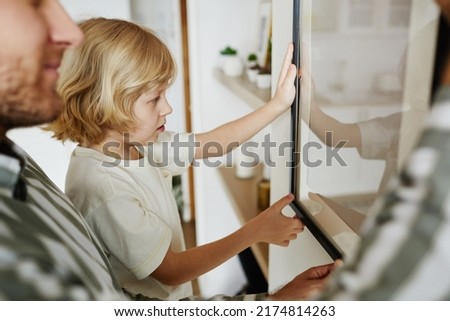 Side view portrait of cute blonde boy helping father hanging picture on wall while decorating new house