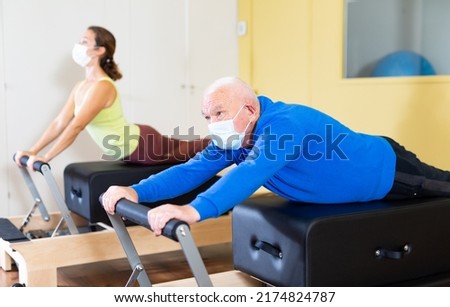 Focused senior man wearing medical mask to protect against viral infections practicing pilates stretching exercises on reformer as part of remedial gymnastics..