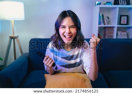 Excited young Asian woman watching TV and cheering sport games competition with laugh victory on sofa at home. Royalty-Free Stock Photo #2174856125