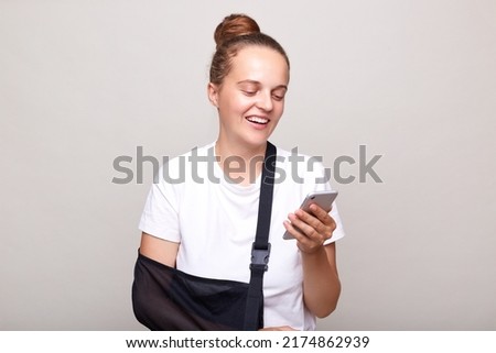 Horizontal shot of positive woman with bun hairstyle wearing T-shirt standing in arm sling, injured her elbow, using mobile phone for finding clinic address, posing isolated on light gray background.