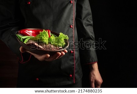 The chef is holding a plate with chopped meat and vegetables. Food preparation concept on dark background