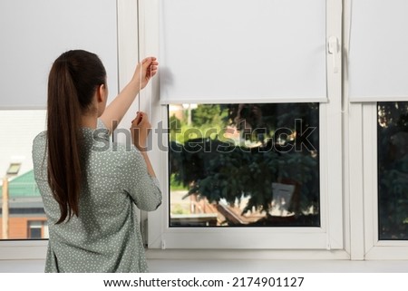 Woman opening white roller blind on window indoors, back view Royalty-Free Stock Photo #2174901127