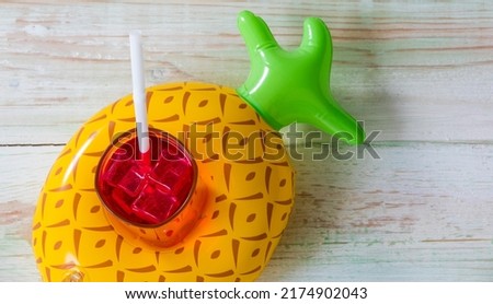 Closeup studio top view shot colorful pineapple shape rubber ring floating holder with iced cold drink glass with straw, sunglasses, small fan and weaving rattan hat with string hatband on wood table. Royalty-Free Stock Photo #2174902043