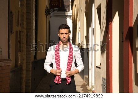 Portrait of young Spanish man in white shirt and black pants dancing flamenco with a red handkerchief with white polka dots in a very narrow street. Typical Spanish concept, dance, culture, tradition.
