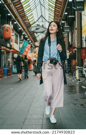 full length with vertical shot of relaxed asian female traveler with camera looking into distance while exploring inside local kuromon ichiba market in Osaka japan